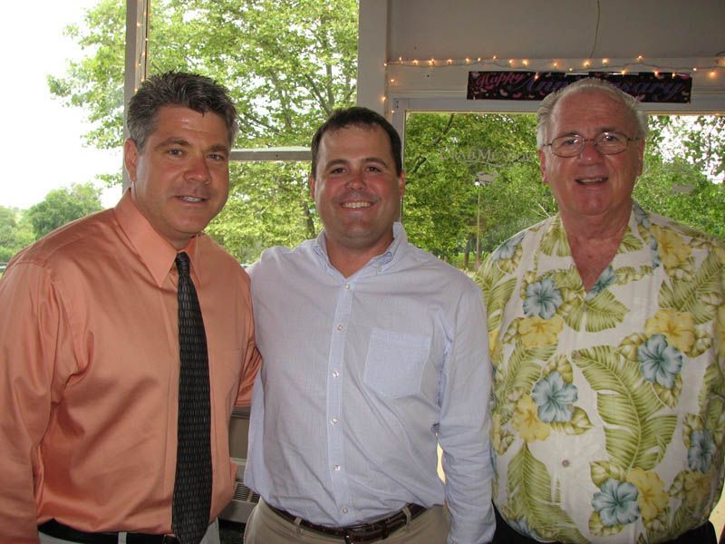 Three men smiling, standing together outdoors. One in an orange shirt, one in a light blue shirt, one in a floral shirt.