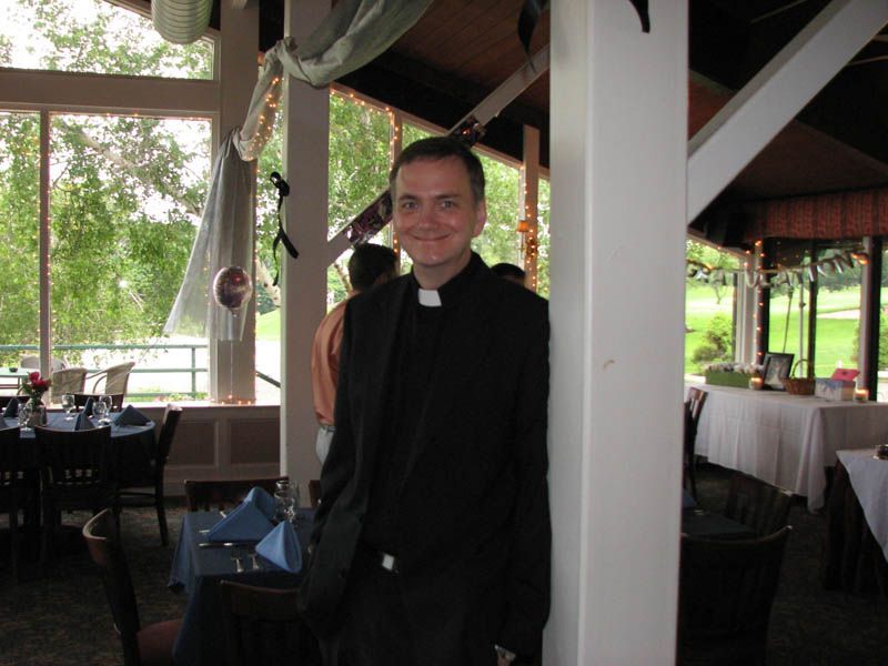 Man in clerical collar and black suit leans against a white pillar in a restaurant.