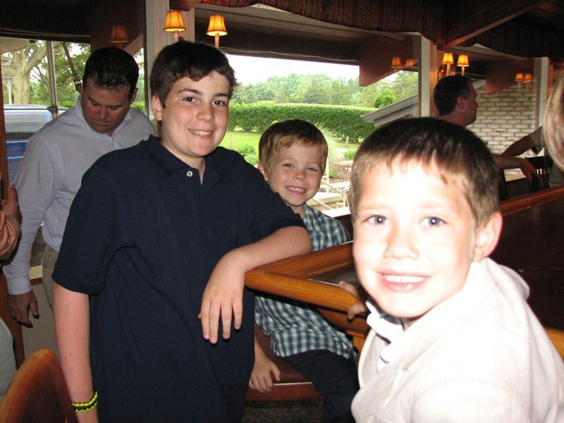 Three smiling boys at a bar, leaning on it. An adult in a white shirt and others are in the background.