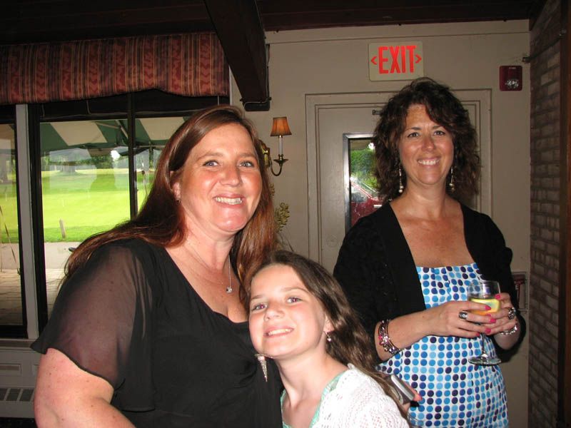 Three smiling people indoors: a young girl with two women, one holding a drink.