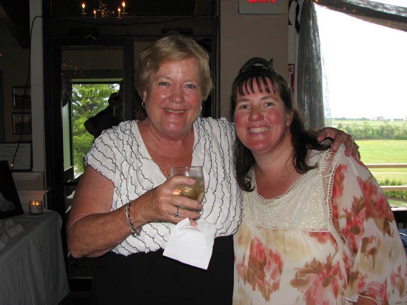 Two women smiling, one holding drink; inside near a window overlooking field.