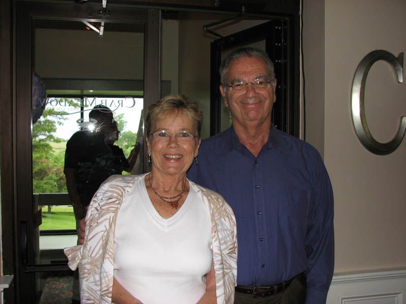 Smiling couple posing indoors; woman in white top, man in blue shirt. 