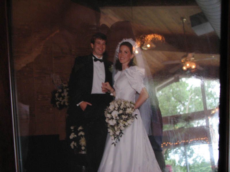 Wedding portrait: couple in formal attire, bride holding bouquet, groom in tuxedo. Set in a venue with windows.
