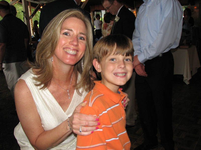 Woman in white dress smiles, arm around a boy in orange shirt; both smile at camera. Inside, with blurred background.