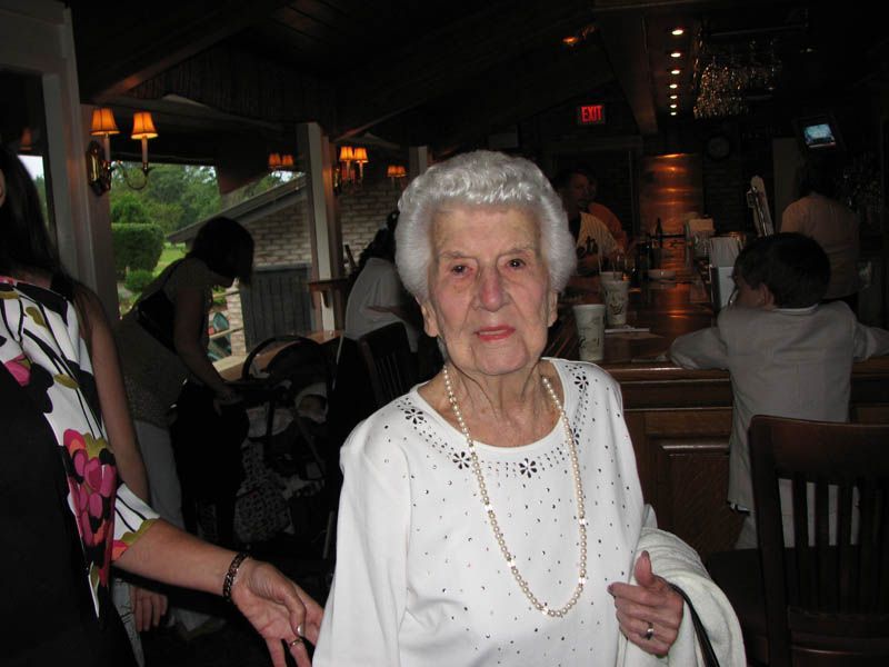 Elderly woman in a white sweater, pearl necklace, smiling at the camera in a restaurant.
