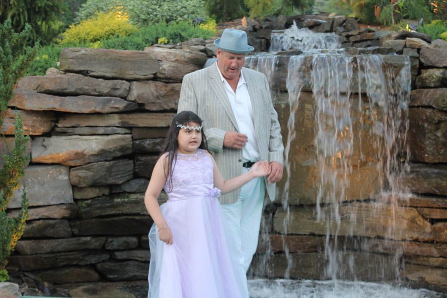Man in light suit and girl in lavender dress stand near a small waterfall feature.