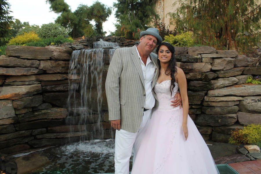 Man in a suit and woman in a pink dress pose by a small waterfall.