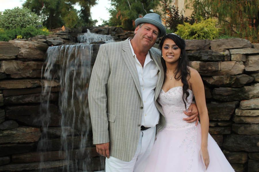 Man in blazer and fedora with young woman in pink dress by a waterfall.
