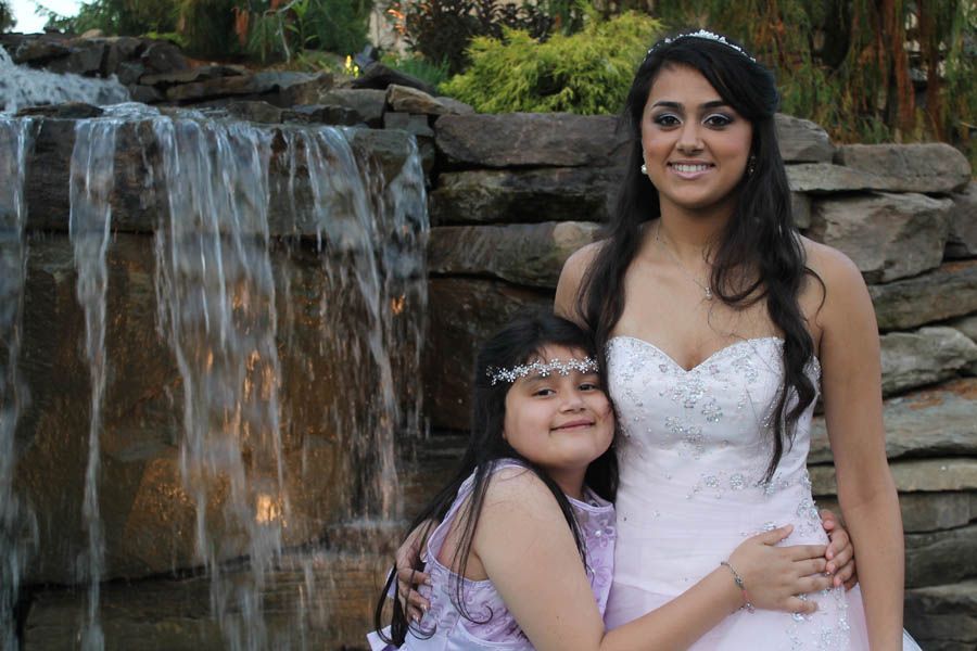Two smiling women, one in a white strapless gown, other in a purple dress, posing near a waterfall.