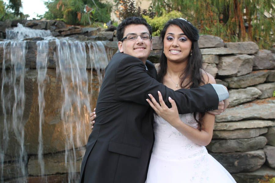 Couple hugging in front of a waterfall; woman in white dress, man in a suit, both smiling.