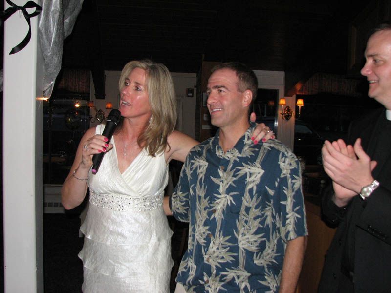 Woman in white dress speaks into microphone, arm around man in Hawaiian shirt; third man applauds.