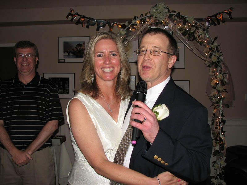 Couple embraces; man speaks into microphone. Party setting with floral arch and man in striped shirt.