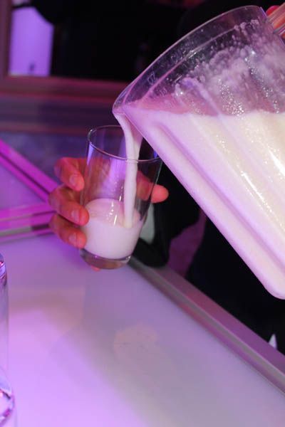 A hand pours a thick, white liquid from a pitcher into a glass, on a white surface with pink light.