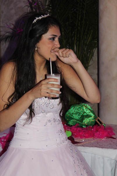 Woman in a pink dress, wearing a tiara, drinking from a glass with a straw, biting a fingernail.