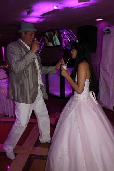 Man in light suit and hat dances with a bride in a white gown, both holding drinks with straws in a party setting.