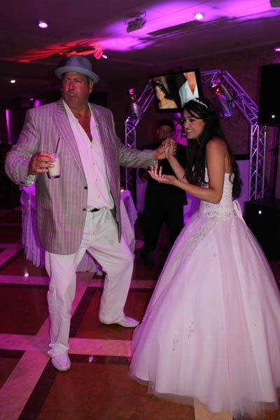 Man in white suit and hat dances with a woman in a white wedding dress. Pink lighting.