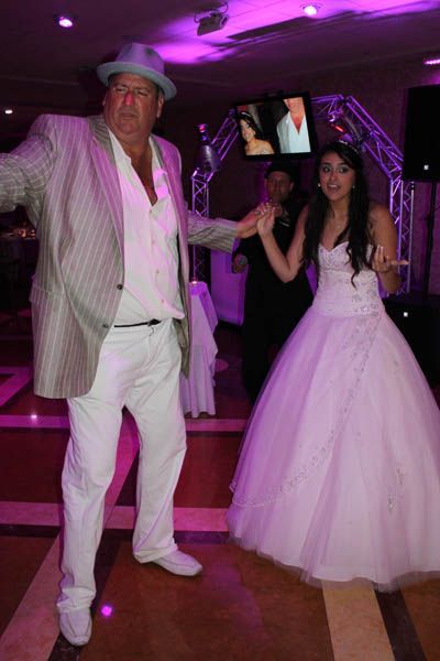 Man in white suit and hat dances with a woman in a white gown at a party. Pink and purple lighting.