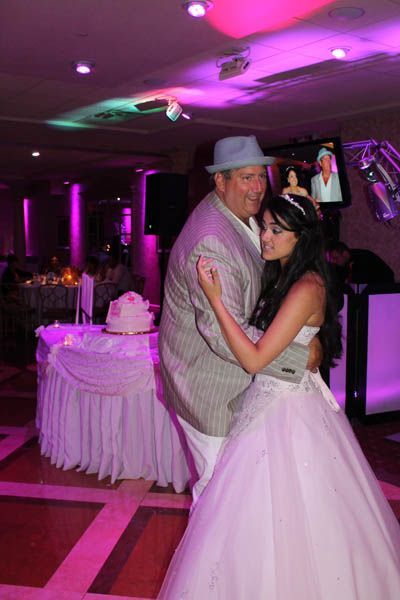 Man in hat dances with person in white gown at a party with a cake.