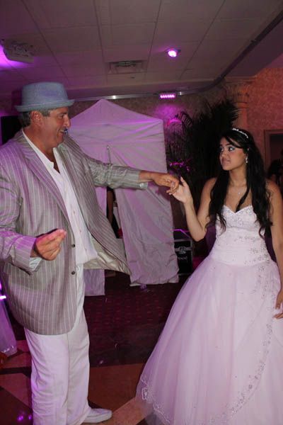 Man in a fedora dances with a girl in a white gown at a party, holding hands, purple lighting.