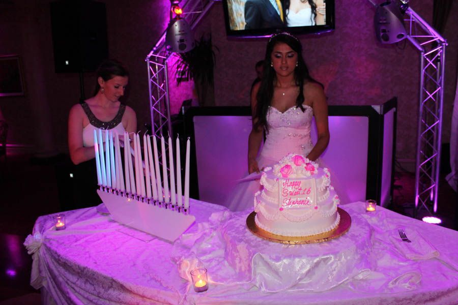 Teen in white dress with cake at a celebration, lit candles, purple lighting, white table.