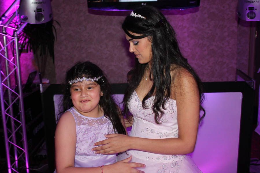 Two women in formal dresses at a party, one touching the other's chest, purple lighting.