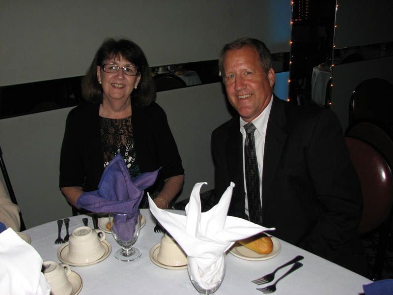 Two people at a table; woman in black jacket and man in suit, white tablecloth, folded napkins.