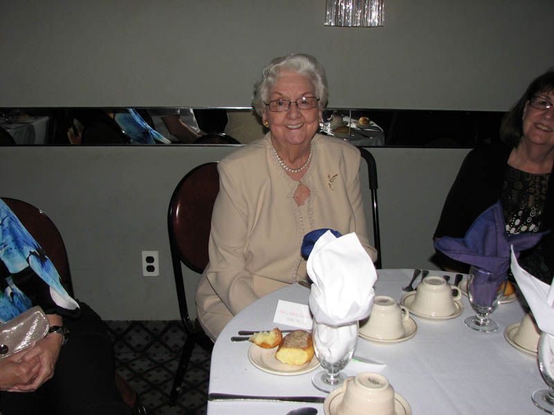 Woman seated at a table smiling, wearing a light-colored suit jacket, a necklace, and glasses.