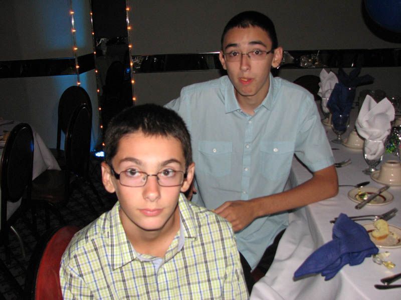Two boys seated at a banquet table. One in a plaid shirt, the other in a light blue button-down, both wearing glasses.