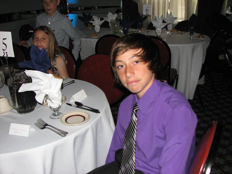 Teen in purple shirt and tie at a table, looking at the camera. Other people and tables in background.