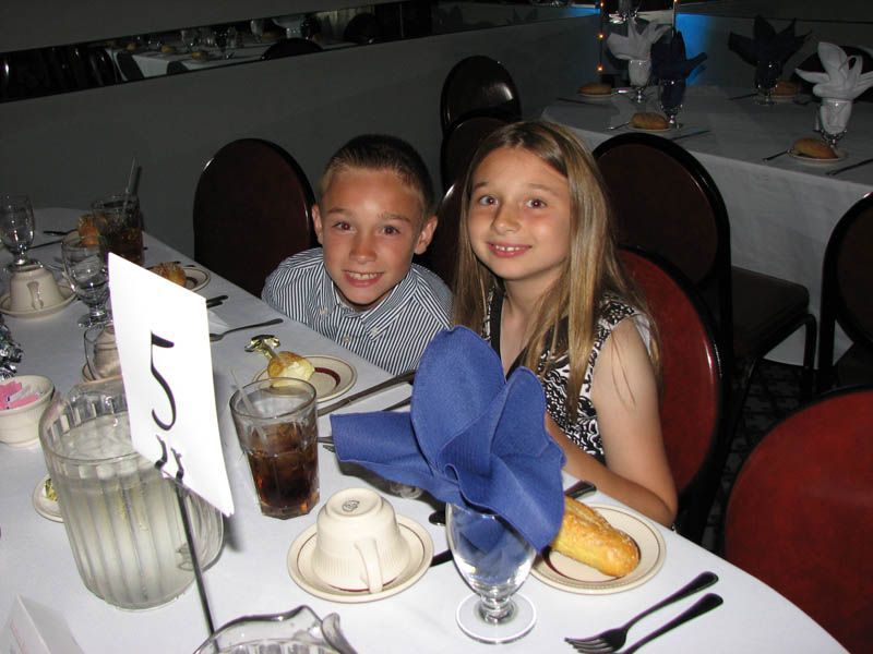 Two children at a table, smiling at the camera. They are at a formal event, with white tablecloths and place settings.