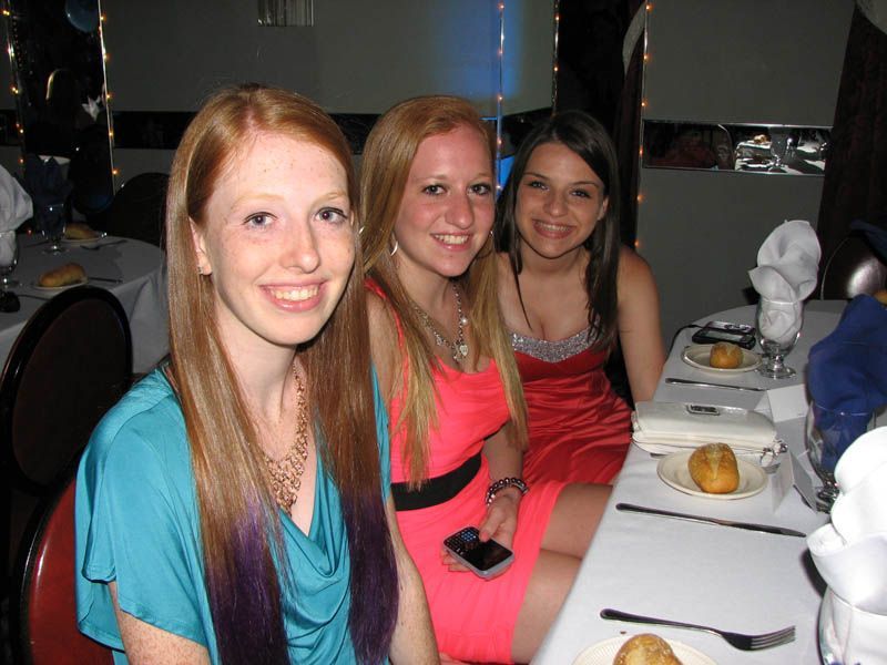 Three smiling people at a table, two in bright dresses. Indoors, festive setting with white tablecloths.