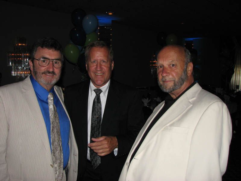 Three men in suits pose indoors with balloons in the background.