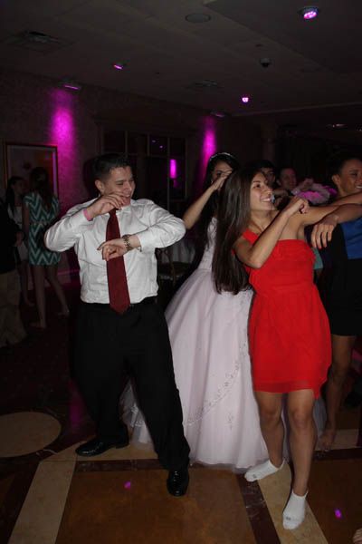 People dancing at a wedding reception. Woman in red dress, man in white shirt, bride in the middle. Pink lighting.
