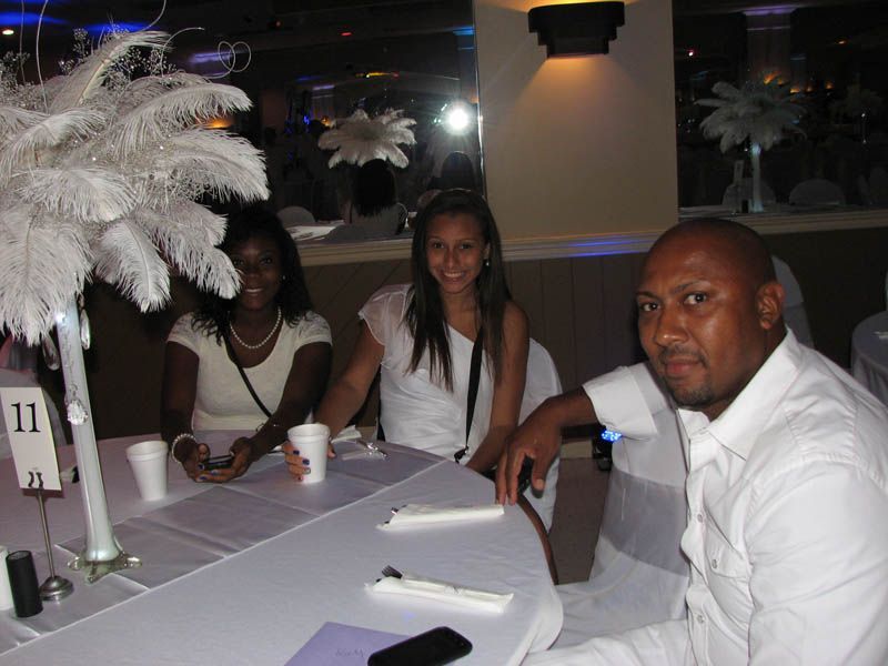 Three people at a table, two women and a man, at an event. White attire. White feather centerpiece.
