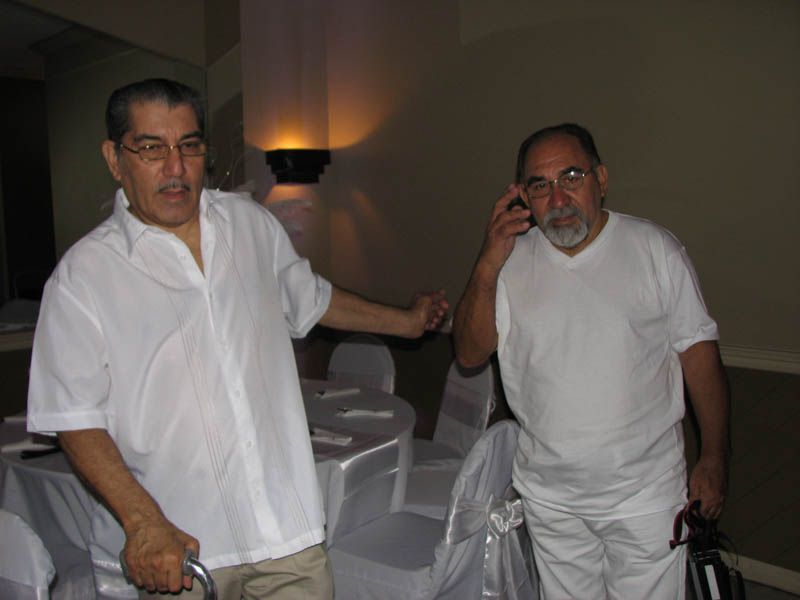Two men in white shirts, one gesturing, the other on a phone, in a room with tables and chairs.