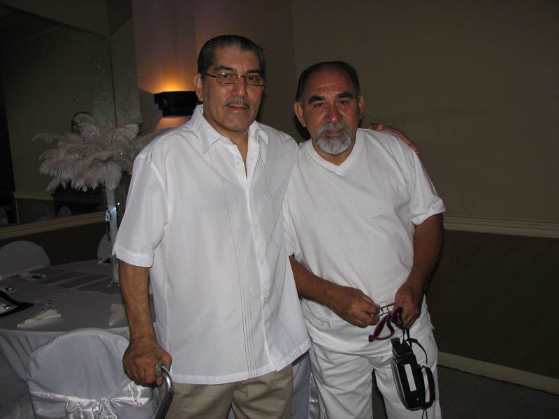 Two men in white shirts pose indoors, one with glasses holding a cane, the other with a beard.