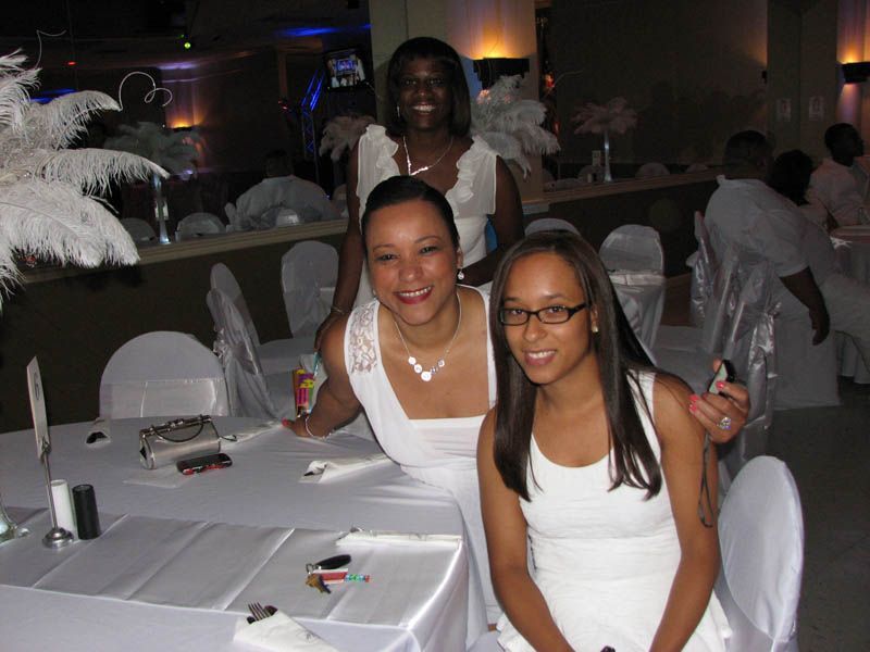 Three people at a white-themed event, smiling. One in front, two behind. Setting is a banquet hall.