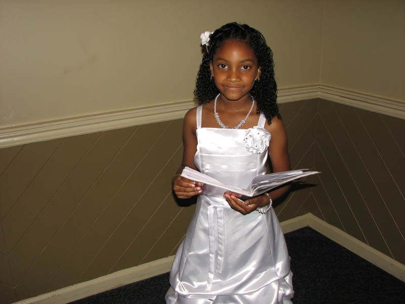 Girl in white dress holding papers, standing against a neutral-colored wall, smiling.