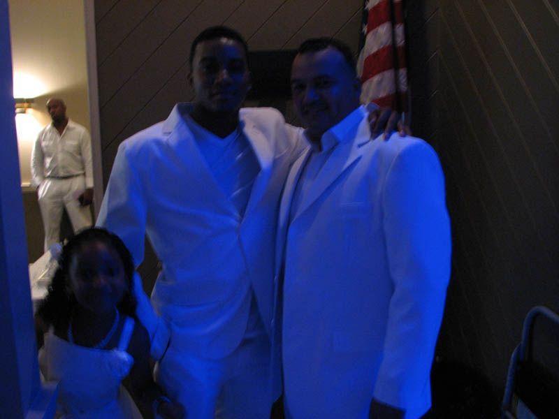 Three people in white suits posing indoors; one with a child. Blue lighting.