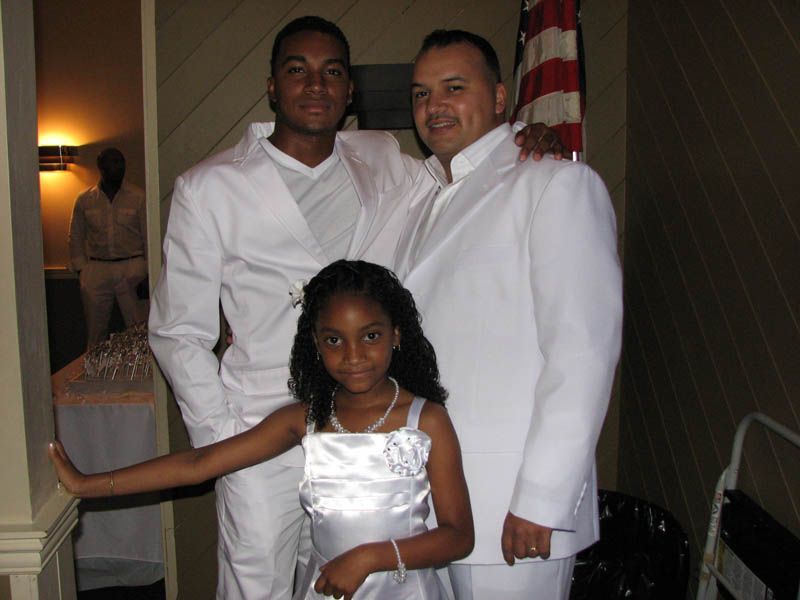 Three people in white formal wear posing for a photo indoors. An American flag is in the background.