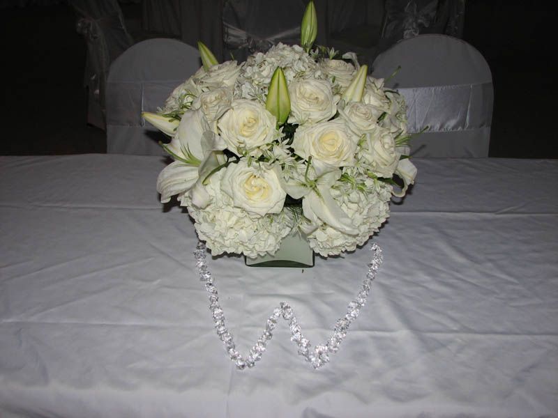 White floral centerpiece with roses, lilies, and hydrangeas on a white tablecloth; two chairs in background.