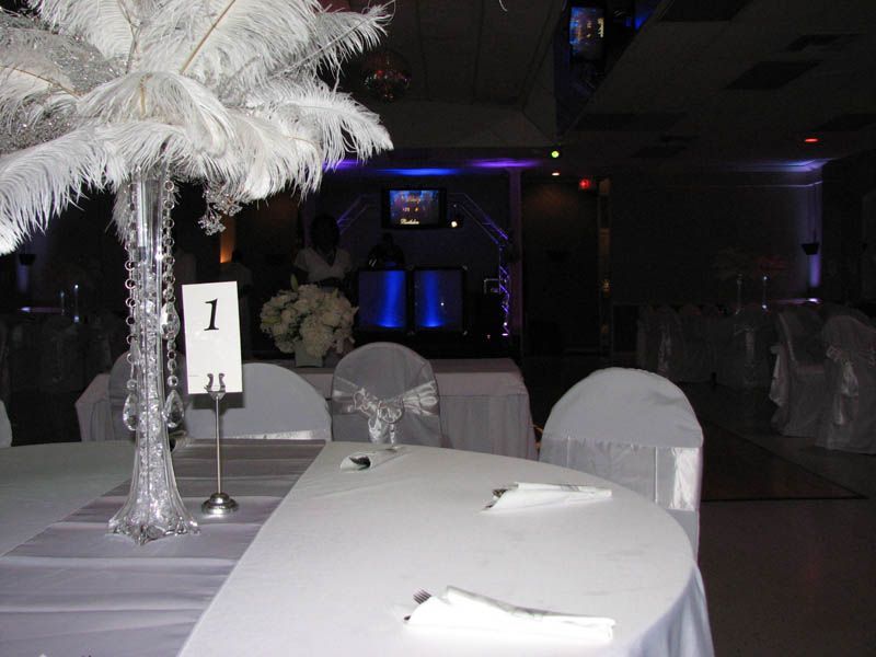 A decorated banquet hall, round tables set with white linens, centerpiece with white feathers, table number 1.