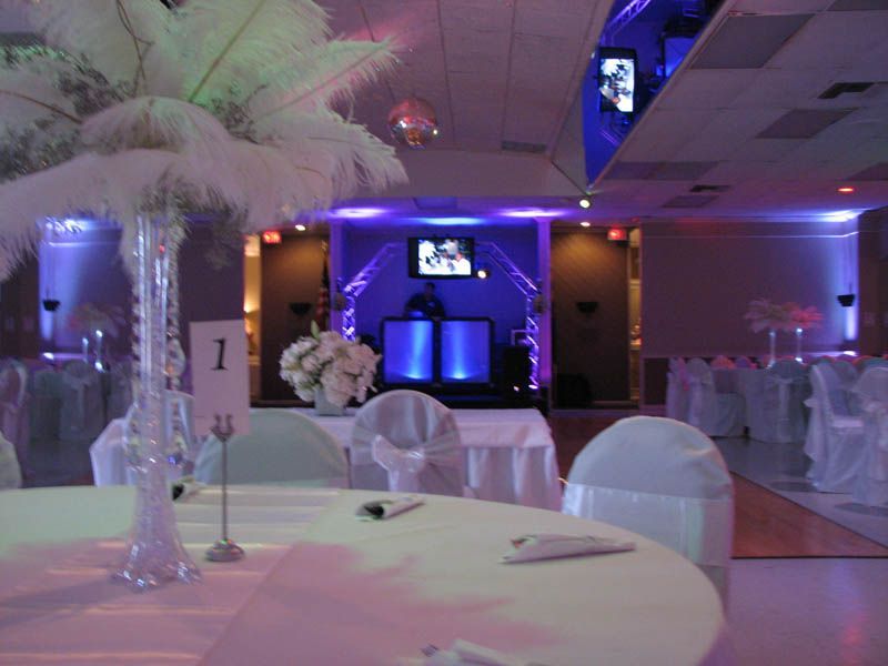 Elegant banquet hall decorated in white with feather centerpieces, tables set, and DJ stage with blue lighting.