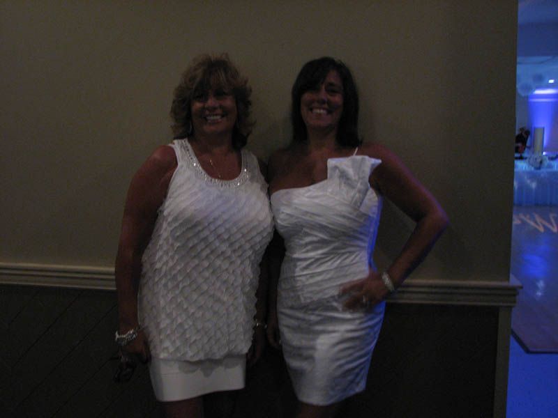 Two women smile, pose in white dresses against a beige wall, possibly at a party.