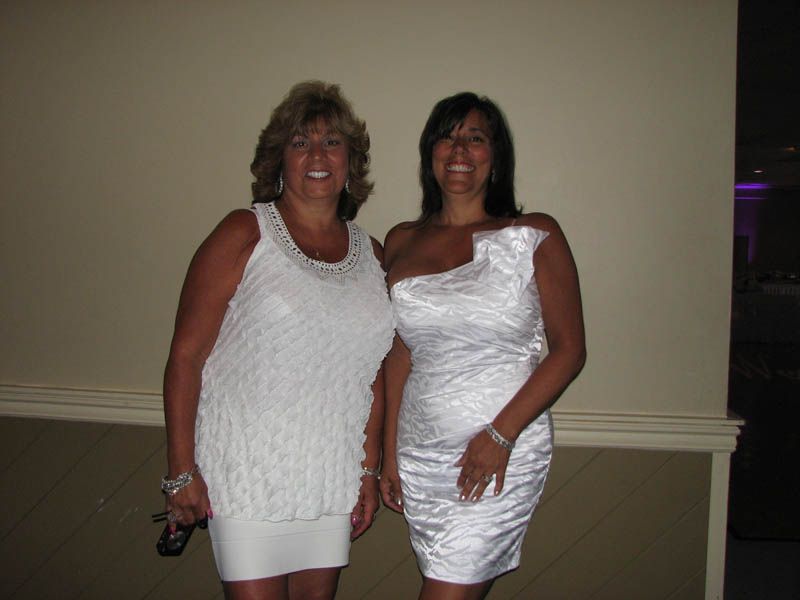 Two women in white dresses smiling in front of a neutral-colored wall.