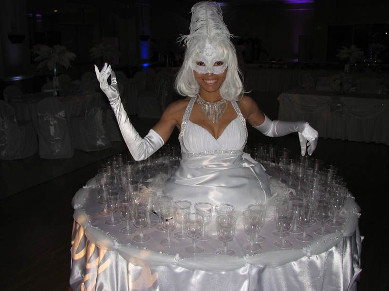 Woman in white dress with champagne glasses on skirt-like table, wearing mask and gloves.