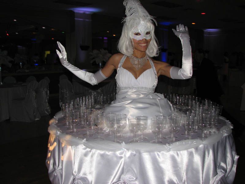 Woman in white dress with champagne glasses, wearing a mask and gloves, at a party.