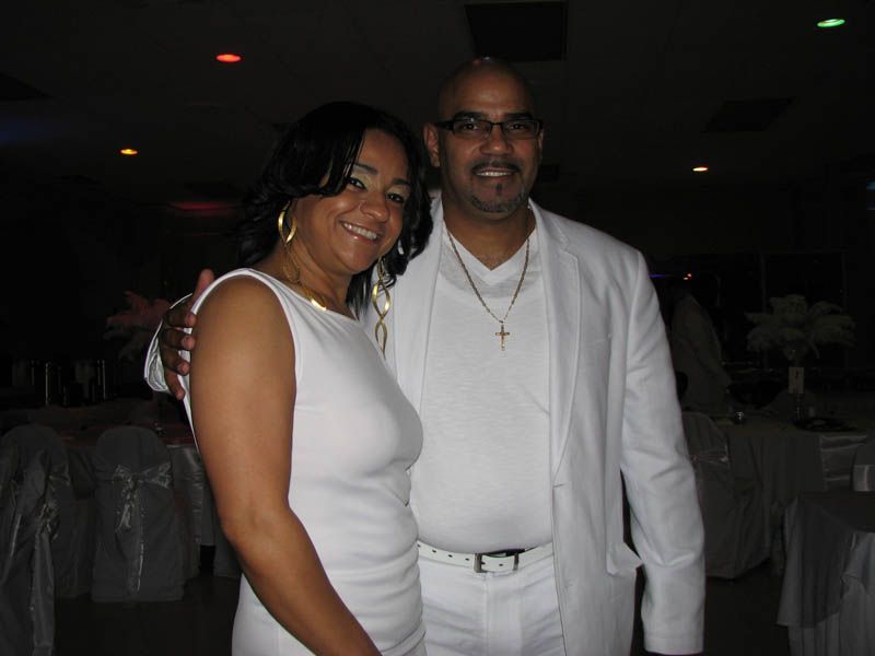 Couple in white outfits smile, posing together indoors at an event.