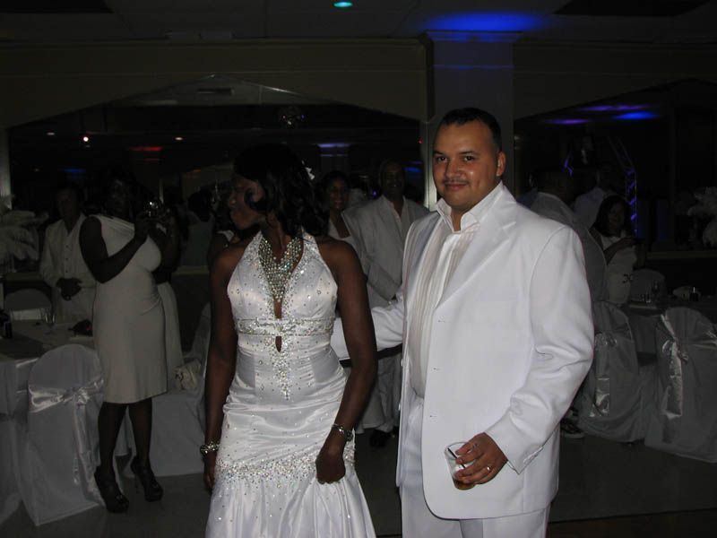 Couple in white outfits at a celebration, likely a wedding. Guests in white outfits in background.