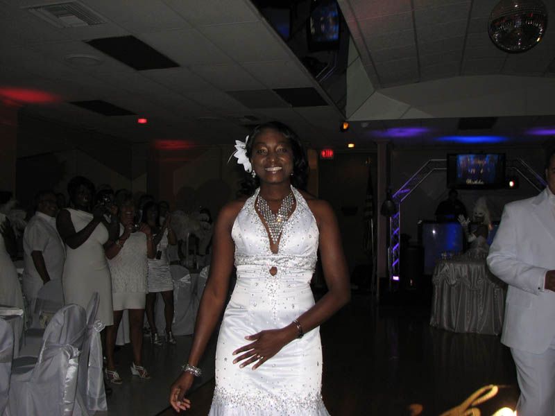 Woman in white dress smiles, walking towards the camera. Guests in white attire applaud in a decorated hall.